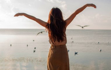 Kolları havada mutlu bir kadın, kumsalda, açık havada uçan kuşlara karşı duruyor.