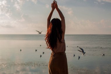 Kolları havada mutlu bir kadın, kumsalda, açık havada uçan kuşlara karşı duruyor.