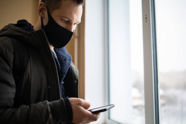 Kafkas 'lı genç adam cep telefonunu pencerenin yanında dururken maske takıyor.