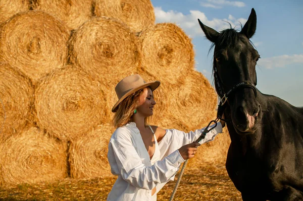 夏の田舎で茶色の馬を撫でる白いシャツの若いブロンドの女性 — ストック写真