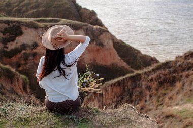 Mavi Deniz 'in arka planında Şapkalı Genç Sıska Kadın Uçurumda Oturuyor.