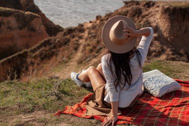 Mavi Deniz 'in arka planında Şapkalı Genç Sıska Kadın Uçurumda Oturuyor.