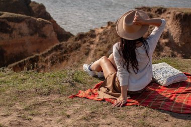 Mavi Deniz 'in arka planında Şapkalı Genç Sıska Kadın Uçurumda Oturuyor.