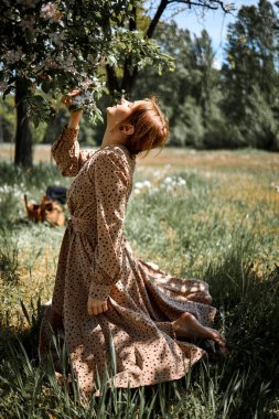 Pembe elbiseli, beyaz, genç bir kadın çiçek açan elma ağacının yanında duruyor..