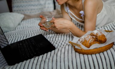 Yatakta ayçöreği ve kahve içerken dizüstü bilgisayar kullanan güzel bir kadın.