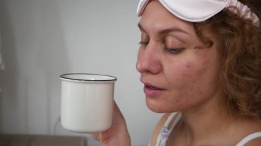 Sarışın kadın bir fincan çay içer ve sabahın erken saatlerinde yatağında telefona bakar.