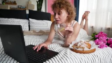 Yatakta ayçöreği ve kahve içerken dizüstü bilgisayar kullanan güzel bir kadın.