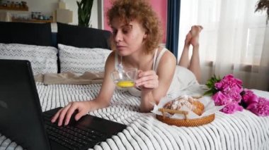 Yatakta ayçöreği ve kahve içerken dizüstü bilgisayar kullanan güzel bir kadın.