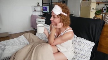 Sarışın kadın bir fincan çay içer ve sabahın erken saatlerinde yatağında telefona bakar.