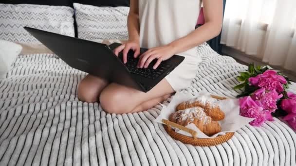 漂亮的女人一边喝咖啡一边用笔记本电脑 床上放着羊角面包 — 图库视频影像