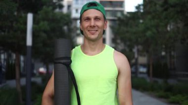 Portrait Of Smiling Young Man In Sport Clothes With Sport Mat In Hands After Training Outdoor.
