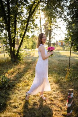 A Woman At The Park Wearing A Sweet Cute Dress Holding Flowers On A Warm Summer Day At Sunset.