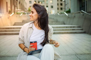 Güzel bir iş kadını, ofis binalarının yanında kahve içerek çalışacak. Başarılı bir iş kadınının portresi. Elinde bir bardak sıcak içecekle şehir sokaklarında işe giderken..