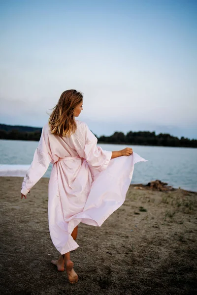 日落时分 一位穿着粉色长袍的浪漫的金发女子在海边 — 图库照片