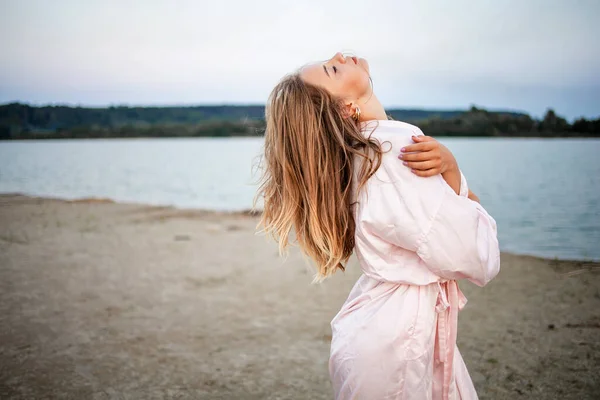 日落时分 一位穿着粉色长袍的浪漫的金发女子在海边 — 图库照片
