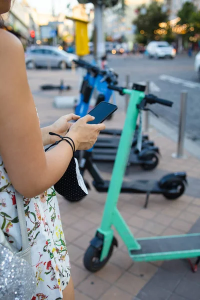 Woman\'s hands close up holding a phone near the modern city electric kick scooter, going to unlock it. Modern city transportation.