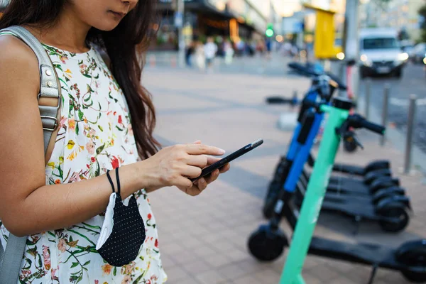 Woman\'s hands close up holding a phone near the modern city electric kick scooter, going to unlock it. Modern city transportation.