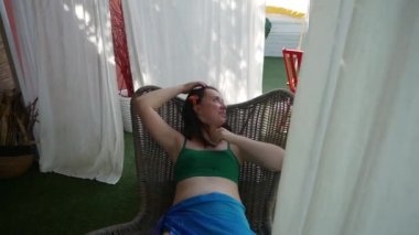 Beyaz kadın dışarıdaki terasta hasır sandalyede oturuyor. Gezgin blogcu kız Bali köyünde tatilin tadını çıkarıyor