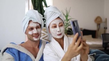 Evde gülümseyen ırklar arası bir çift. Güzellik maskeli ve akıllı telefonlu mutlu Asyalı kadın ve yatak odasında dinlenen beyaz adam. Evlilik hayatı cep telefonu kullanan insanlar için selfie çekmek..