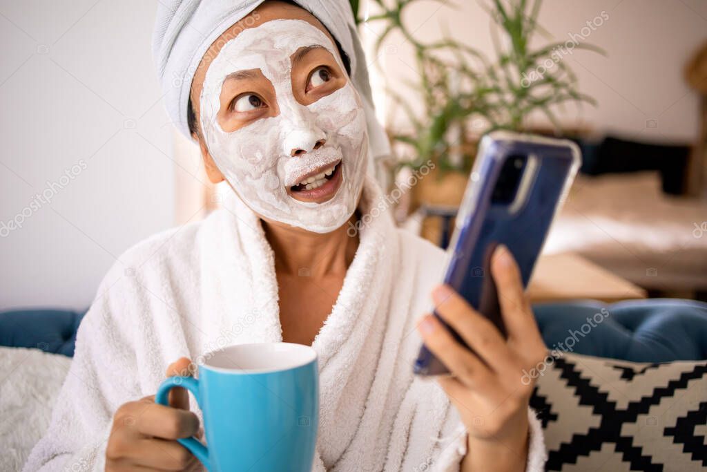 Happy surprised shocked asian woman apply facial mask and using cellphone got a good news when sitting on the couch at home. Beauty skincare and wellness morning concept.