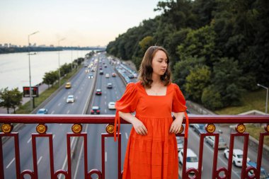 Kızıl saçlı, mavi gözlü, turuncu elbiseli beyaz kadın köprüde duruyor..