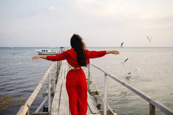 Asian Girl in red standing with open arms on wooden bridge against sky, seagulls fly over the beach. Freedom concept