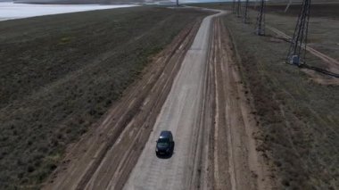 Siyah araba rüzgar türbinlerinin yakınındaki bir yolda ilerliyor. İHA ya da kuadrokopterden ateş ediliyor. 