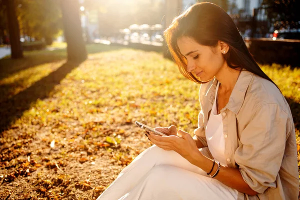 Sonbaharda gün batımında parkta çimlerin üzerinde dinlenen cep telefonu kullanan genç bir kadın..