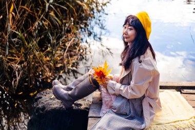 Sıcak botlar giyen ve elinde sonbahar yapraklarıyla gölün kıyısındaki ahşap iskelede oturan bir kadın.