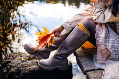 Sıcak botlar giyen ve elinde sonbahar yapraklarıyla gölün kıyısındaki ahşap iskelede oturan bir kadın.