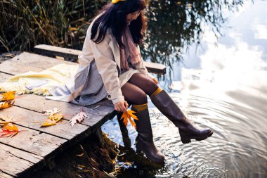 Sıcak botlar giyen ve elinde sonbahar yapraklarıyla gölün kıyısındaki ahşap iskelede oturan bir kadın.