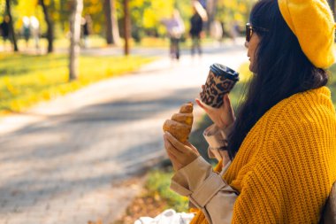 Sonbahar parkında otururken elinde kahve ve kruvasan tutan sarı kazaklı bir kadın..
