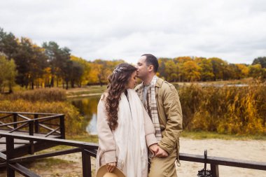 Aşk ve aşk kavramı. Tahta köprüde kucaklaşan ve birbirlerine bakan tatlı genç çift, gölün arka planında sonbahar ağaçları..