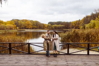 Aşk ve aşk kavramı. Tahta köprüde kucaklaşan ve birbirlerine bakan tatlı genç çift, gölün arka planında sonbahar ağaçları..