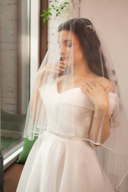 Şık, esmer, klasik beyaz elbiseli gelinin duvak altında yakın çekim pozu. Gelin portresi, makyaj ve saç stili, moda gelini. Peçeli güzel gelin.