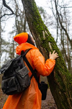 Yeşillik bir ormanda sırt çantalı genç Asyalı bir kadın ağaca sarılıyor. Uzun ağaçlar ve yumuşak orman ışıklarıyla çevrili doğayla barışçıl bir bağ anı.