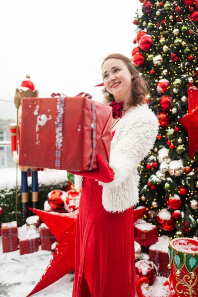 Kış tatili konsepti. Genç bir kadın dışarıda Noel ağacının yanında hediye kutusu tutuyor. Mutlu kadın yeni yılı kutluyor..