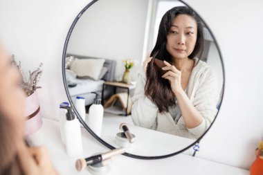 Son sınıf çekici, orta yaşlı, gri saçlı Asyalı kadın ayna yansımasına bakıyor. Karışık gri saçlarını tarıyor. Menopoz reklamından sonra Alopecia saç kaybı önleme tedavisi.