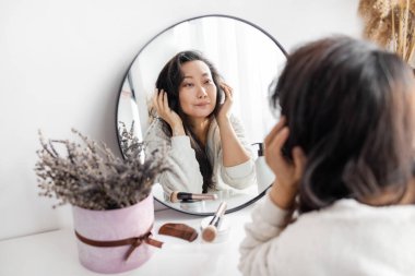 Güzel Asyalı orta yaşlı kadın dışarı çıkmaya hazırlanıyor, sabah makyajını yapıyor ve banyoda aynanın karşısına fırçayla yüz pudrası sürüyor..