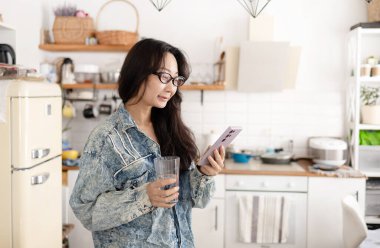 Gülümseyen Asyalı kadın akıllı telefon kullanıyor ve elinde bir bardak su tutuyor..