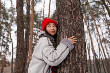 Çekici Asyalı kadın doğa aşığı portresi. Kış boyunca çam ormanında ağaca sarılıyor. Kış ruh hali.