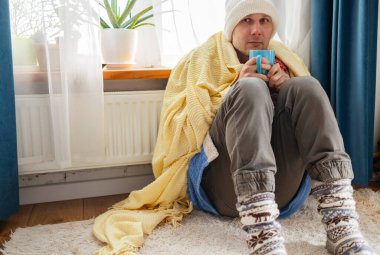 Evdeki soğuk odada, radyatörün önünde şapkası ve atkısı olan beyaz bir adam donuyor. ısıtma sorunları