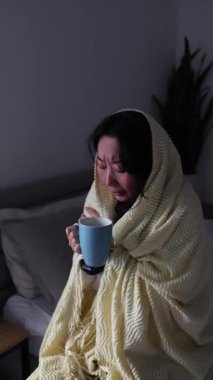 Genç Asyalı kadın sıcak battaniyeyle örtülmüş evde çay içiyor. Kadın soğuktan donar, kötü hisseder, sağlıksız. Grip belirtileri gösteren hasta bir kız battaniyeyle kaplanmış evde sıcak çayla ısınıyor..