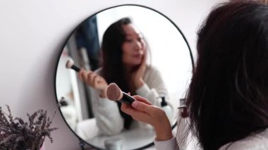 Güzel Asyalı orta yaşlı kadın dışarı çıkmaya hazırlanıyor, sabah makyajını yapıyor ve banyoda aynanın karşısına fırçayla yüz pudrası sürüyor..