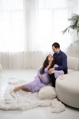 Bebek bekleyen mutlu genç çift. Hamile milenyum kadını ve kocası göbeğine yakın ayakkabılarıyla yatakta oturuyor ve gelecekteki ebeveynliğin tadını çıkarıyorlar..