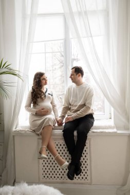 Beyaz tenli çifti seven erkek ve kadın hamileliği parlak pencerenin yanında anarlar.