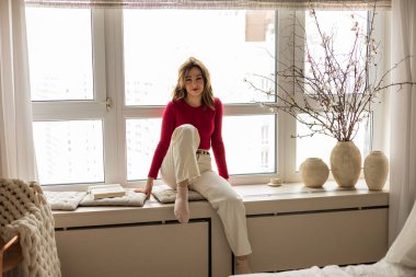 Mutlu, genç, güzel, pozitif, çekici bir kadın pencere eşiğinde oturup kameraya bakarak keyif alıyor. Gülümseyen neşeli bayan evde takılıyor, gülüyor, eğleniyor, resim çiziyor..