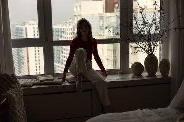 Mutlu, genç, güzel, pozitif, çekici bir kadın pencere eşiğinde oturup kameraya bakarak keyif alıyor. Gülümseyen neşeli bayan evde takılıyor, gülüyor, eğleniyor, resim çiziyor..