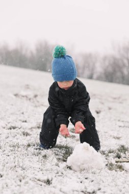 Siyah ceketli bir çocuk kışın arka planında bir kardan adam heykeli yapıyor.