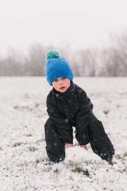 Siyah ceketli bir çocuk kışın arka planında bir kardan adam heykeli yapıyor.